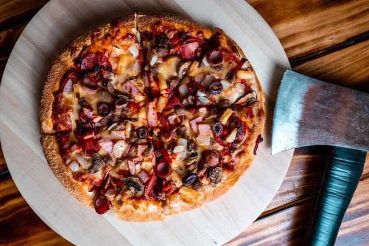 Pizza with meat toppings on a platter beside a cleaver on a wooden surface.