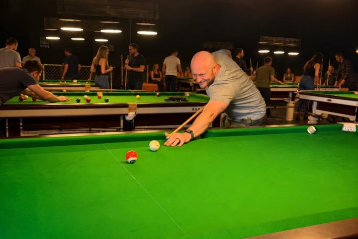 Person playing pool in a dimly lit room with multiple tables.