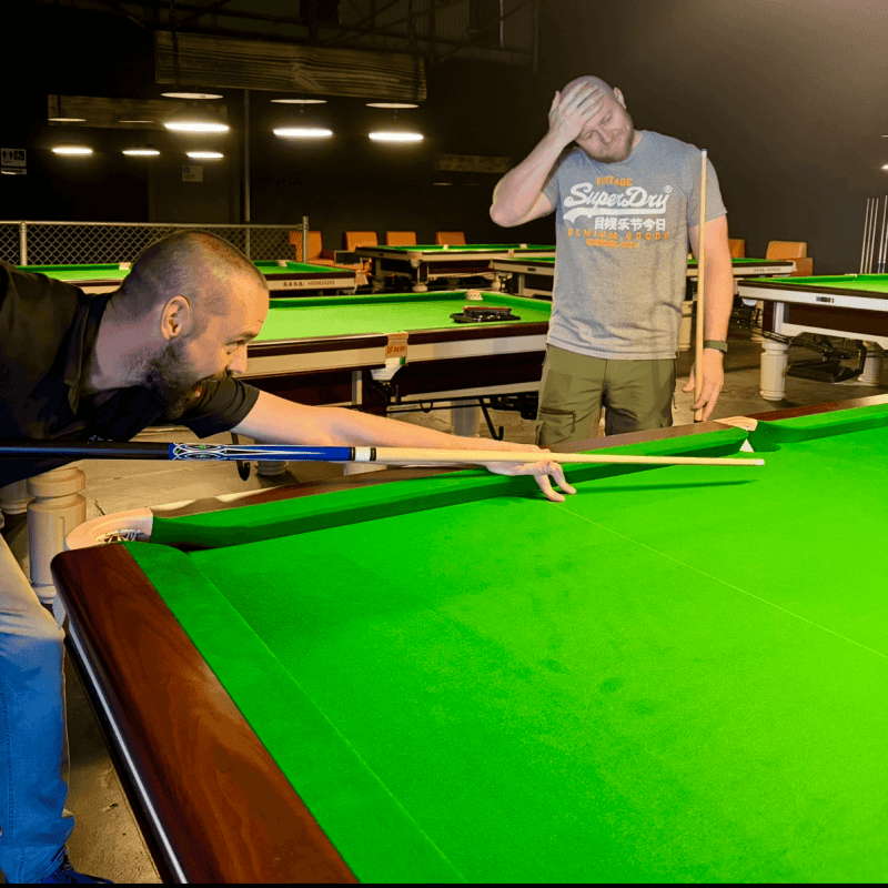 Two men playing billiards, one aiming a shot, the other watching, in a dimly lit room.